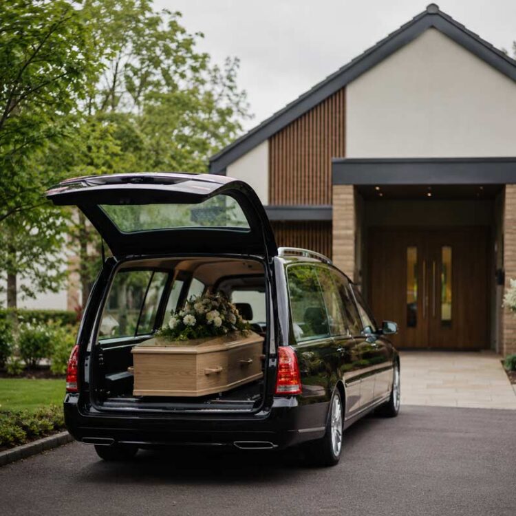 Hearse carrying a simple wooden coffin outside a crematorium with no mourners present.